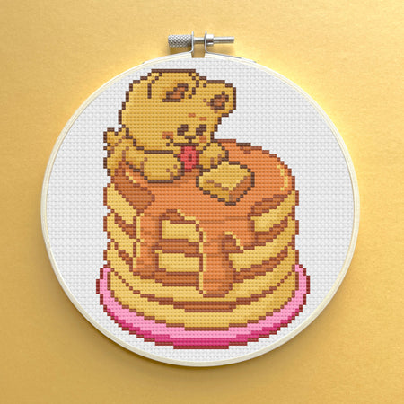Cross-stitched design of a bear on pancakes in an embroidery hoop against a gold background
