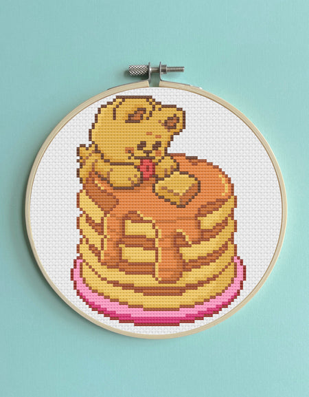 Cross-stitched design of a bear on pancakes in an embroidery hoop against a light blue background