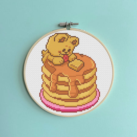 Cross-stitched design of a bear on pancakes in an embroidery hoop against a light blue background