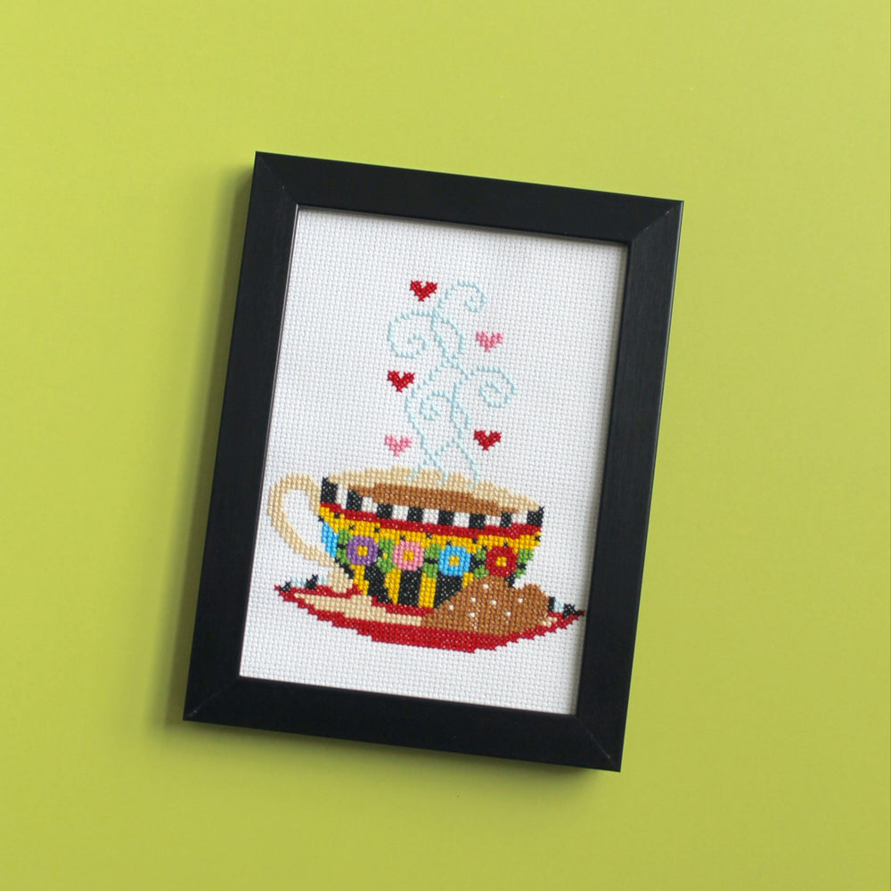 Framed cross-stitched picture of a teacup with hearts on a green wall