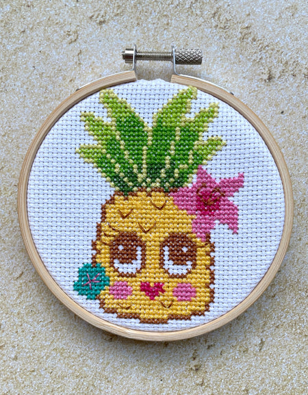 Cross-stitched pineapple with a face and flowers in a wooden hoop on a sandy surface.