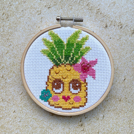 Cross-stitched pineapple with a face and flowers in a wooden hoop on a sandy surface.