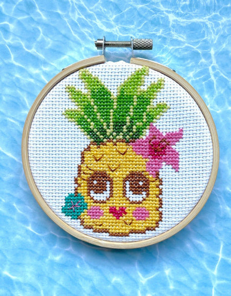 Cross-stitched pineapple with a face in a hoop against a blue water background