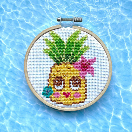 Cross-stitched pineapple with a face in a hoop against a blue water background