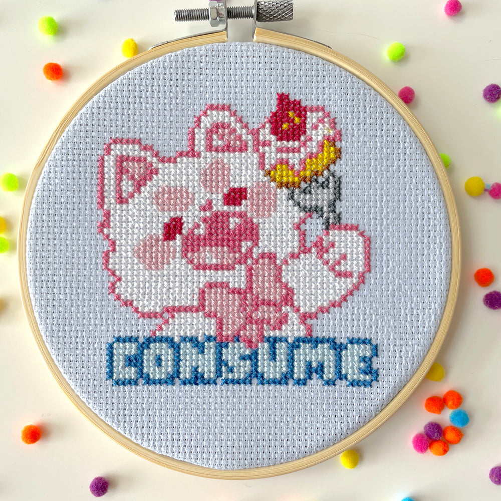 Cross-stitched design of a cartoon character with 'CONSUME' text in an embroidery hoop, surrounded by colorful beads.