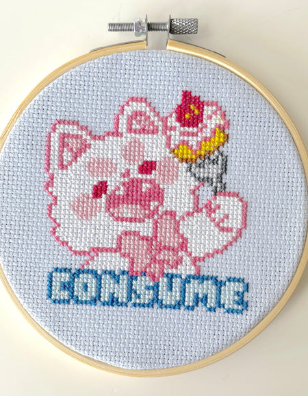 Cross-stitched design of a cartoon character with a cake on its head inside an embroidery hoop.