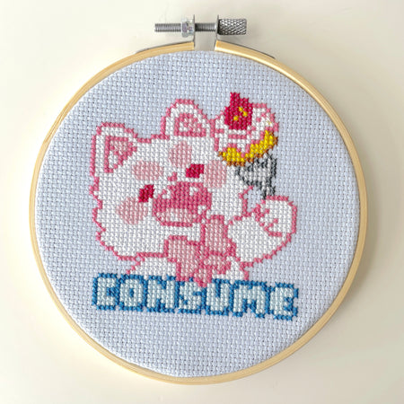 Cross-stitched design of a cartoon character with a cake on its head inside an embroidery hoop.