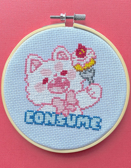 Cross-stitched design of a cat with a and 'CONSUME' text in an embroidery hoop on a pink background.