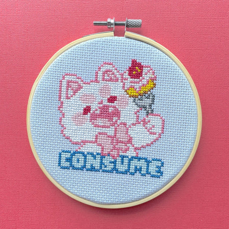 Cross-stitched design of a cat with a and 'CONSUME' text in an embroidery hoop on a pink background.