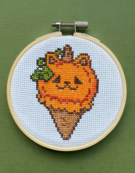 Cross-stitched design of a cat pumpkin ice cream cone with a face in an embroidery hoop on a green background