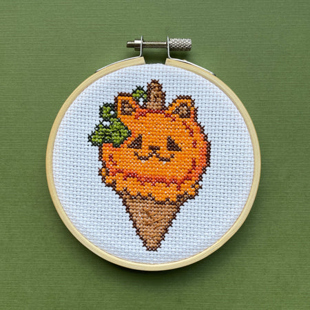 Cross-stitched design of a cat pumpkin ice cream cone with a face in an embroidery hoop on a green background