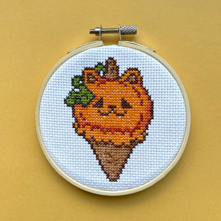 Cross-stitched design of a cat-shaped ice cream cone on a beige background