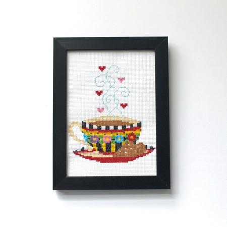 Framed cross-stitched artwork of a teacup with hearts on a white background